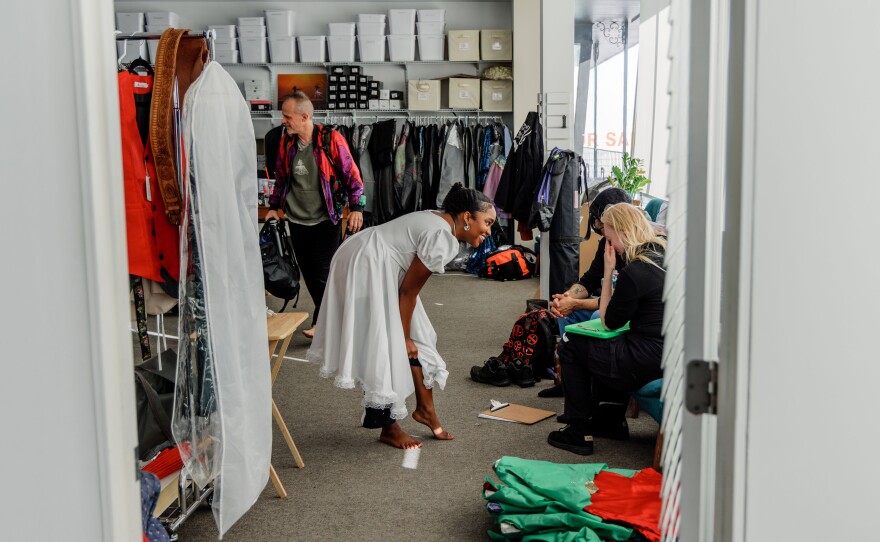 A womanleans over as she is putting on leggings to talk with two people who are seated in a dressing room space. 