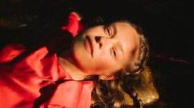 A woman lying down in a darkened space looks directly at the camera.
