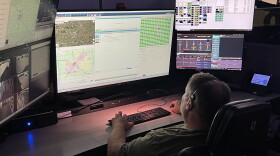 An emergency dispatcher sits in front of a bank of screens with maps and other information displayed.