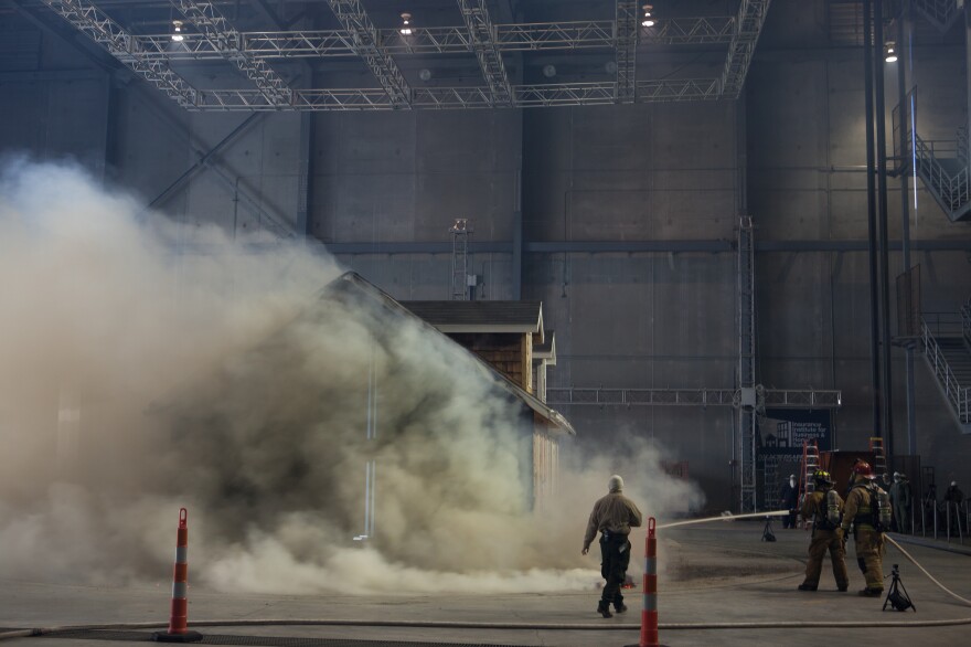 Members of a local fire department work with research facility staff to extinguish the burning test home.