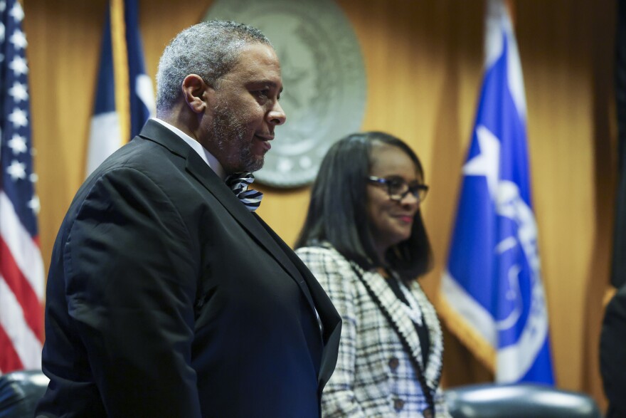 A photo of Tarrant County Commissioner Roderick Miles Jr.,  a Black man with salt-and-pepper hair and a short beard. He's wearing a black suit and a bowtie, looking to his side while standing at the commissioners court dais in front of some flags and the seal of Tarrant County. His colleague Alisa Simmons, a Black woman with shoulder-length straight hair, stands beside him smiling, slightly out of focus.