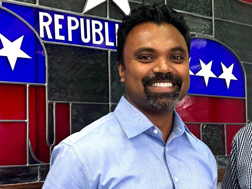 A man with a goatee in a blue button down shirt smiles in front of a stained glass window that says "Republican".