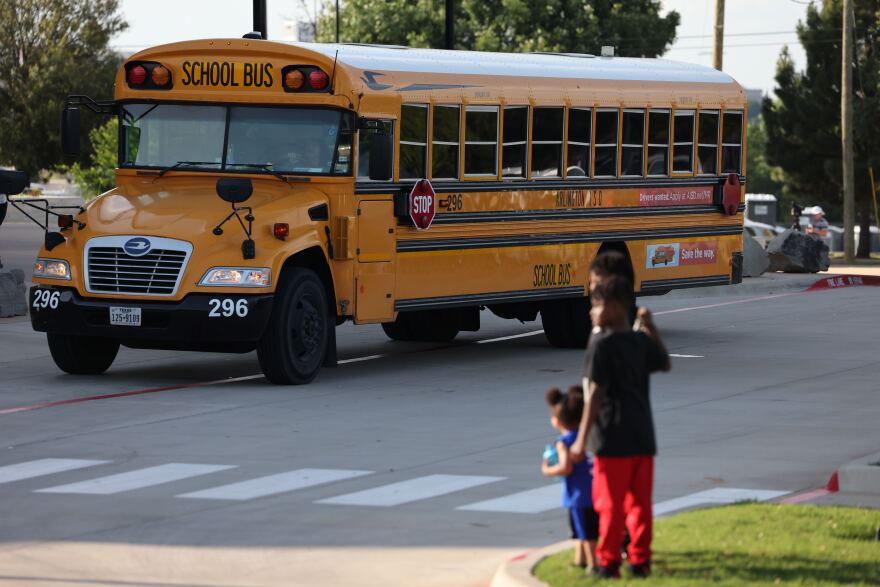 A photo of a yellow school bus with three shildren standing in the foreground.