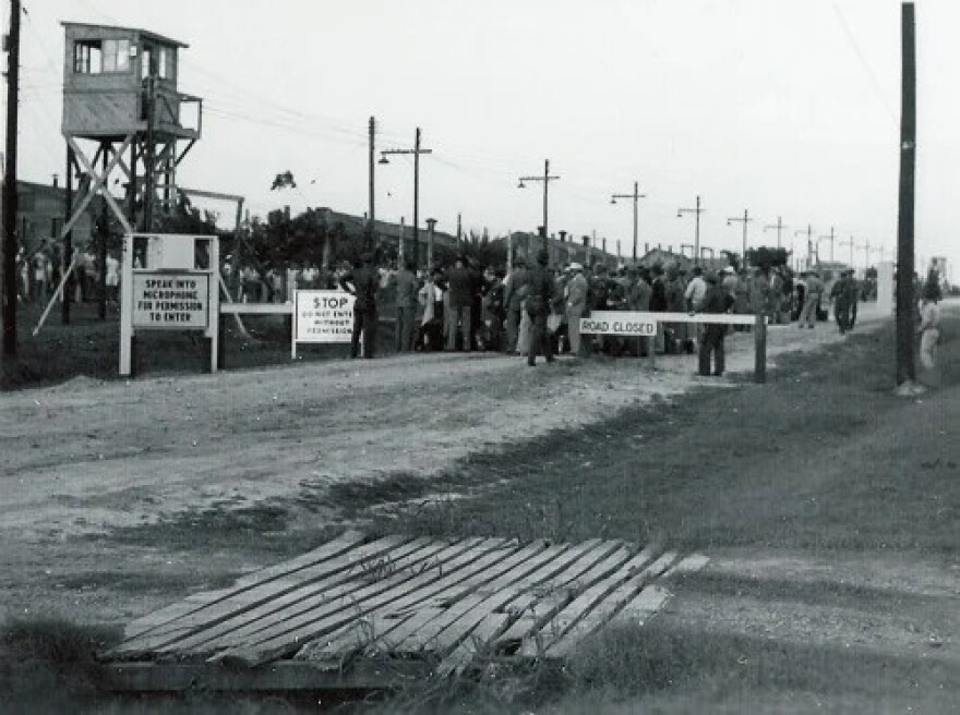 A guard tower and barbed wire fence. There are people behind the fence. In front of the fence there is a road, barred by a road closed sign and guards. The ground is bare.