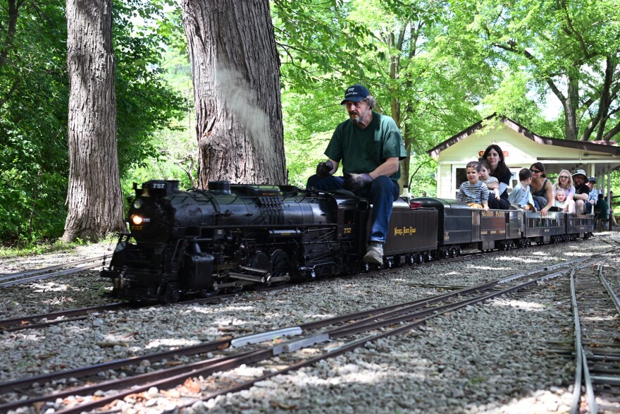 A man drives a miniature steam engine down the tracks with passengers riding behind