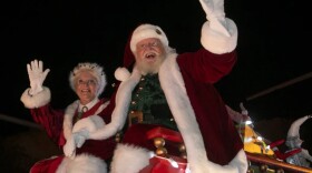 A man dressed as Santa Clause with a red and white coat, glasses, white gloves waving on a parade float with a similarly dressed woman to the left
