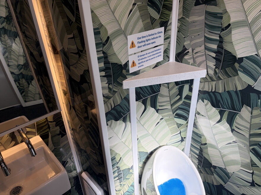 A photograph of the sink and urinal inside the Throne. A sign reads: "Use time is limited to 10 minutes. Flashing lights indicate the door will soon open." 