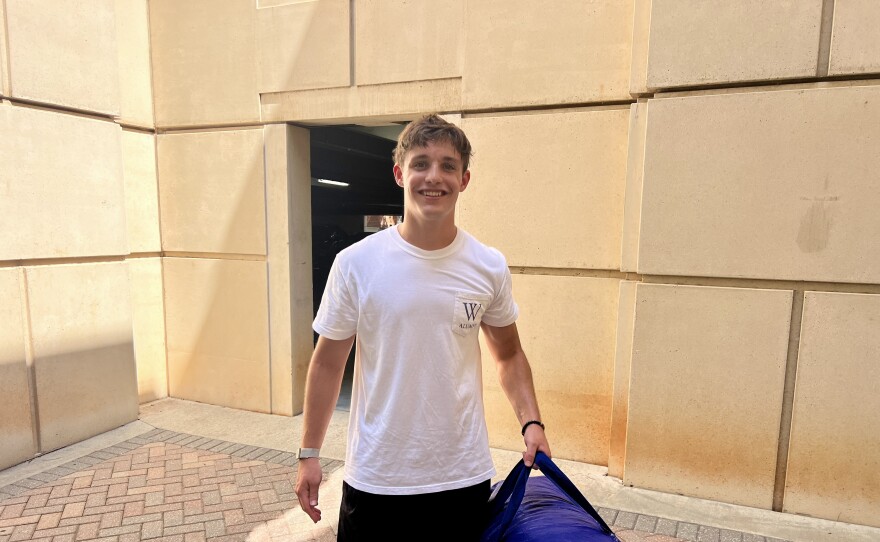 A young man in a white T-shirt smiles while carrying a large blue bag.