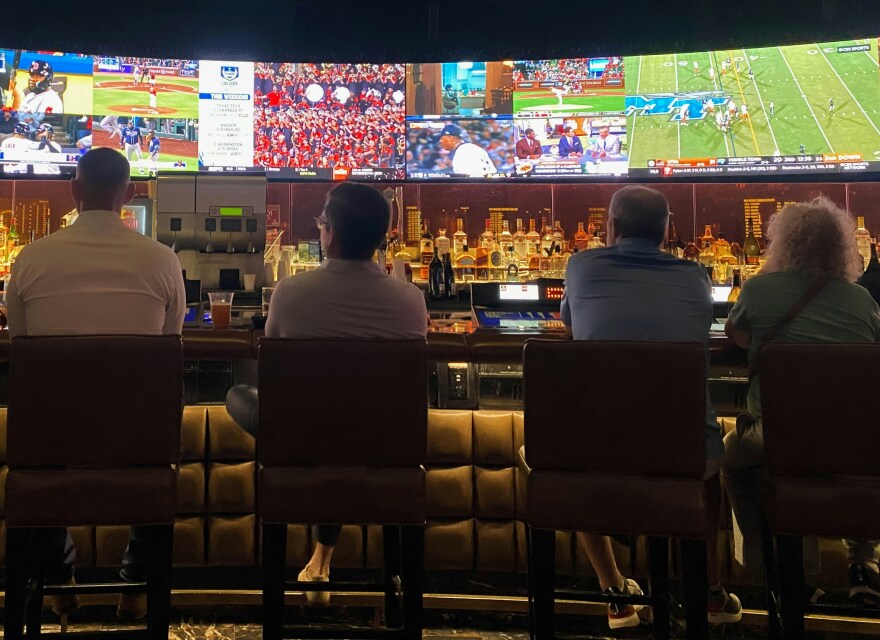 Patrons sit at a sports bar watching matches on four TV screens mounted behind the bar. 