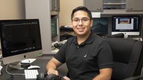 Andrew Méndez is sitting at a desk and looking at the camera and smiling.