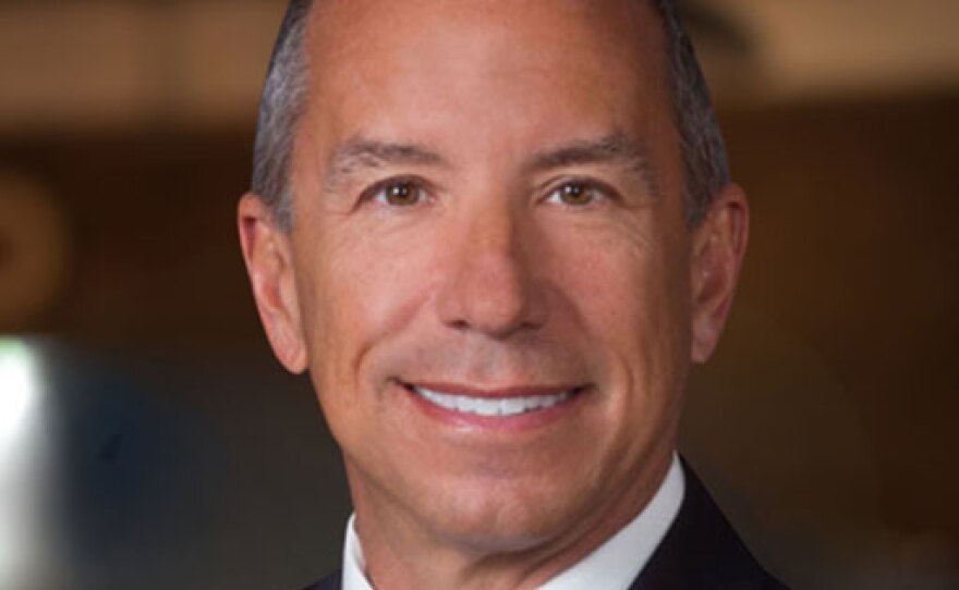A smiling man with short grey hair, wearing a white collared shirt, a gold tie, and a black suit coat