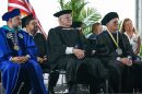Three men in graduation attire sit on chairs on a stage. The American flag is in the background. 