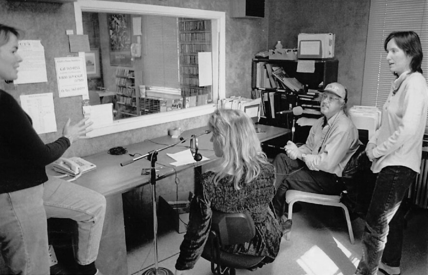 Staff members have a discussion in a radio control room surrounded by microphones and office supplies.
