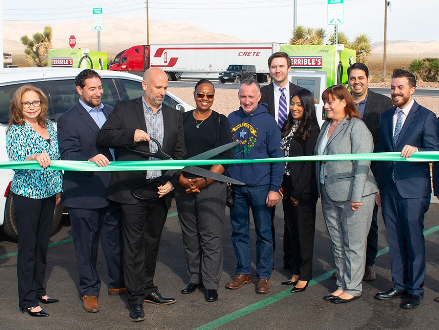 A ribbon-cutting ceremony with 13 people standing in front of a Terrible Herbst EV charging station, with a dry mountain landscape behind them.