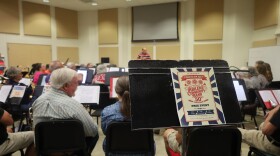 A flyer of the Abilene Community Band's 50th anniversary flyer sits on a music stand, as the orchestra prepares for rehearsal in the background.