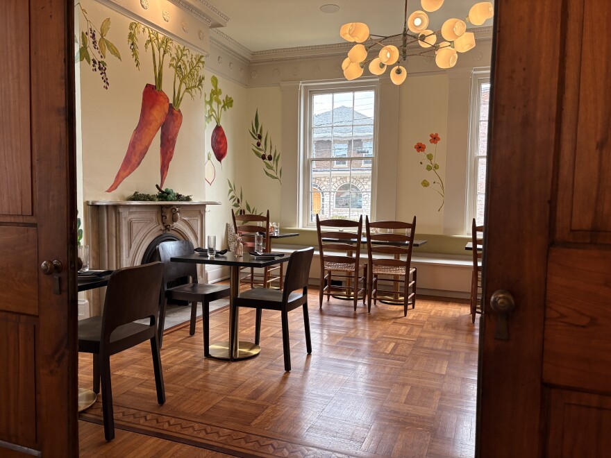 Two open doors frame a dining room with wooden floors, a fireplace, and vegetables painted on the walls.