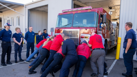 A group of around 9 people push a fire engine into its bay. Most of them are wearing red shirts. They are surrounded by onlookers wearing navy shirts. A man sits in the driver's seat of the fire engine. 