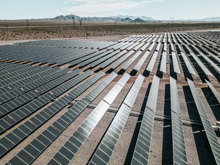 A large solar farm in the desert with mountains in the far background.