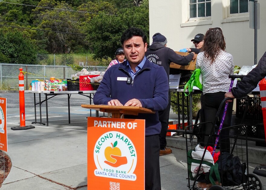 Fernando Cardenas stands at a podium in front of tables of food outside a building.