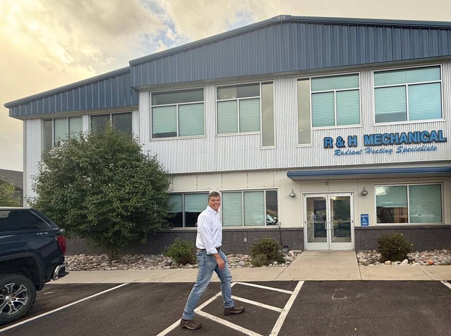 A middle aged man in a white button-down shirt and jeans walks across a parking lot in front of a metal-sided building with a sign that reads "R & H Mechanical" in blue letters. 