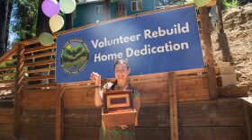 A woman holds a wooden jewelry box and a house key in front of a blue house in the redwoods. Behind her is a banner that reads, "Volunteer Rebuild Home Dedication"