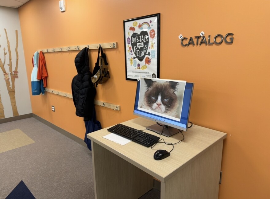 Against a bright orange wall, a desk sits with a desktop computer on it. The computer has a photo of a grumpy cat taped to the screen. Above the computer is a sign that says "Catalog", except the "CAT" has quotation marks around it so it reads like cat-alog.