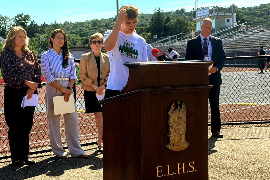 Ryan Dueker, an East Lyme High School lacrosse player and student ambassador at LAX2LIVE, a program to prevent youth suicide, spoke of the stressors that student athletes face on September 8, 2026.. He is flanked by state mental health officials. Behind him in a brown jacket is Ann Dagle, co-founder of the Brian Dagle Foundation and mother of Brian, a former student at the high school, who died by suicide when in college. The foundation runs the LAX2LIVE initiative.