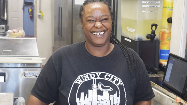 Woman smiling and standing next to a restaurant kitchen and wearing a black shirt with a skyline logo and "Windy City" inscribed on the front.