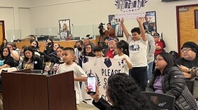 An elementary student speaks at a podium as classmates hold up signs in support of Winston behind him.