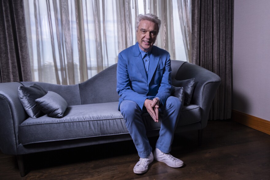 a silver-haired man in a blue suit and white shoes sits on a blue-gray couch