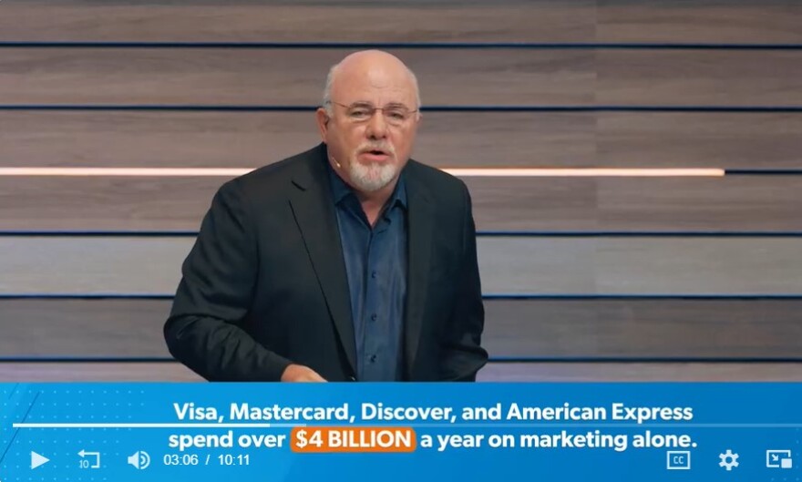 Dave Ramsey gives a talk on stage in front of a wood paneled background, wearing navy blazer