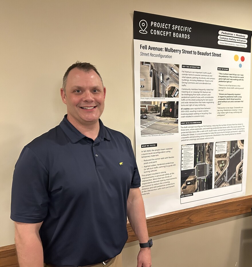 A man in a dark grey polo stands next to a poster board on a white wall and smiles at the camera.