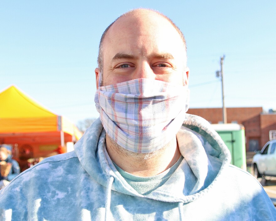 Dale Amann is wearing a plaid face mask, a speckled gray hoodie and smiles into the camera. 