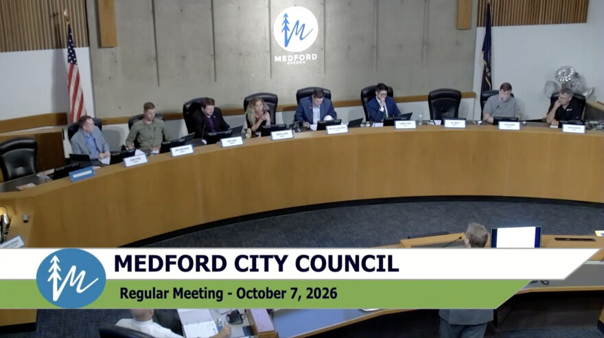 Eight people are seated at a wooden dais. A blond woman in the middle speaks into a microphone. 
