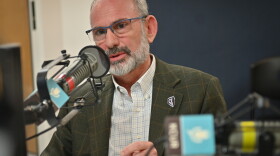 A man sits inside a radio studio. He is talking at a microphone and gesturing with his left hand.