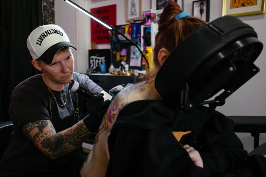 A person lays face down in a massage chair as another person tattoos their back