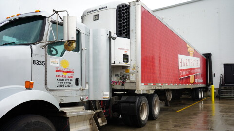 A large semi truck is parked at a warehouse. The trailer is completely red with a large Harvesters logo pasted in the middle of it's face.