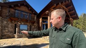 A man motions toward a log cabin.