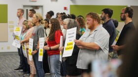 Over a dozen people hold signs in a meeting room. The nearest sign reads, "There is NO substitute for a building substitute." 