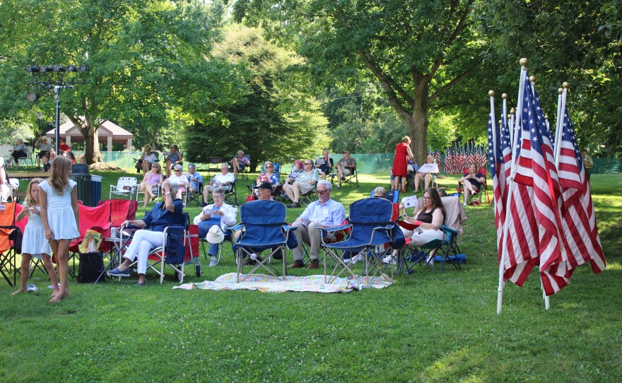 Two young girls stand near lawn chairs as a crowd gathers on the grass at Connie Link Amphitheatre, with rows of American flags visible on the right and a park shelter in the background.