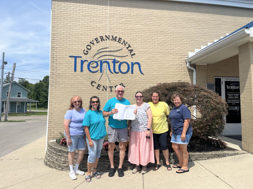 six people stand in front of a brick building that says Trenton Government Center. They're holding a stack of papers