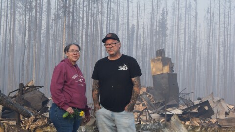 Robert Nelson and Helena Fix-Nelson stand the burned wreckage of their home in Tok on August 19, 2026. Helena holds a marigold and a zucchini from the family’s greenhouse, which survived.