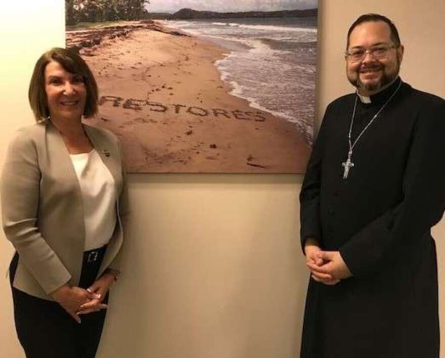 Dr. Deborah Beidel and Father José Rodríguez stand in front of a picture of the word "Restores" written on a beach in Puerto Rico. Photo: Danielle Prieur