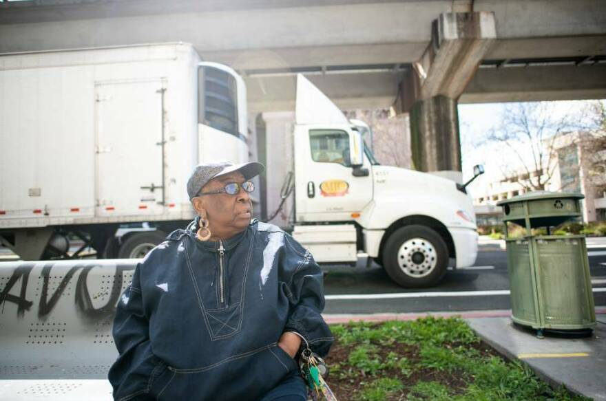 Community activist Margaret Gordon sits on a bench in West Oakland with the BART tracks behind her on March 4, 2022, as a semi-truck stops on 7th Street, on a popular trucking route to the nearby Port of Oakland.