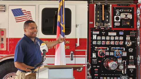 A man gestures to a fire truck while standing at a lecturn.
