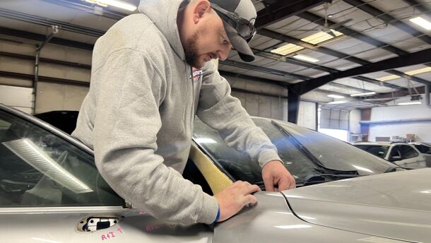 A man wearing a grey hoodie is seen leaning on the front of a car from outside the passenger seat. 