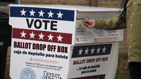 A voter drops off their ballot at a ballot drop box outside that has red white and blue coloring.