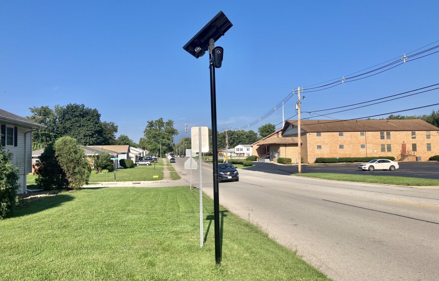 A solar-powered camera mounted on a tall black pole stands beside a suburban street with houses, lawns, and cars on a sunny day.