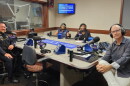 Four smiling people sit around a table in a radio talk studio: a man front left has short light brown hair and a light brown mustache and beard and is wearing a police uniform; a woman back left has short blue hair and is wearing glasses and a blue top; a woman back center has long black hair and is wearing a black top and blazer; a man at right has very short dark hair and is wearing glasses, a grey button-down shirt, and white linen pants