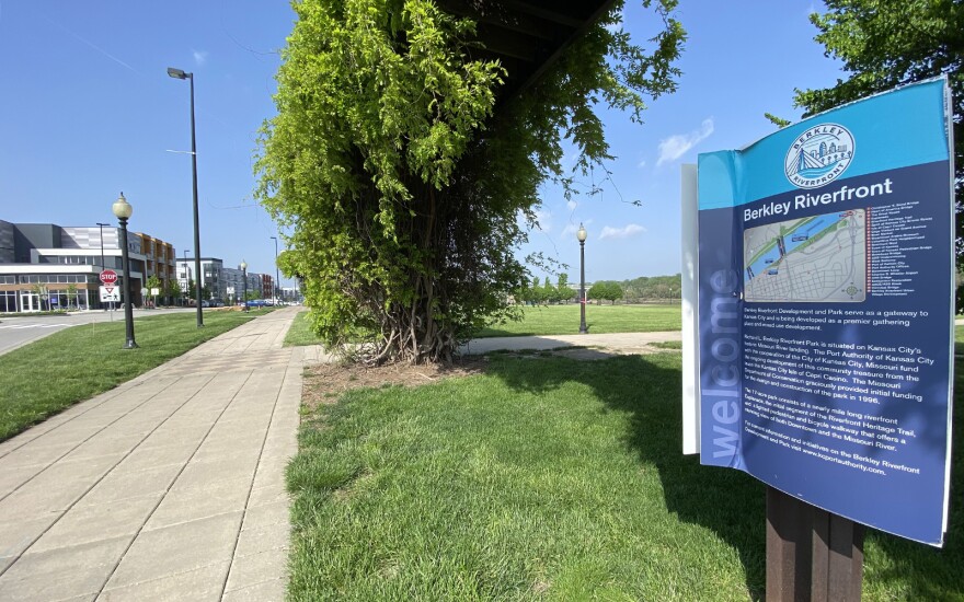 A large blue sign sits at the right of the photo. It's labeled "Berkley Riverfront" and has a map and some information about the park. In the background is a long sidewalk next to a vine-covered trellis and apartment complexes.